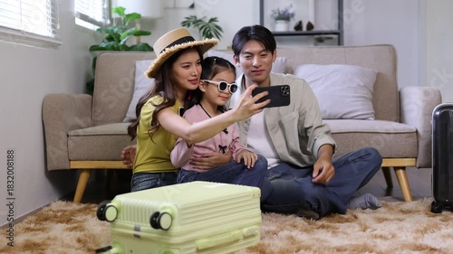 Asian family with their daughter taking a selfie with a smartphone in their living room, getting ready for a holiday trip with their packed suitcases and smiling together happily.