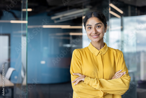 Portrait of a young Indian businesswoman in a yellow shirt with her arms crossed on her chest standing in the office and looking confidently at the camera