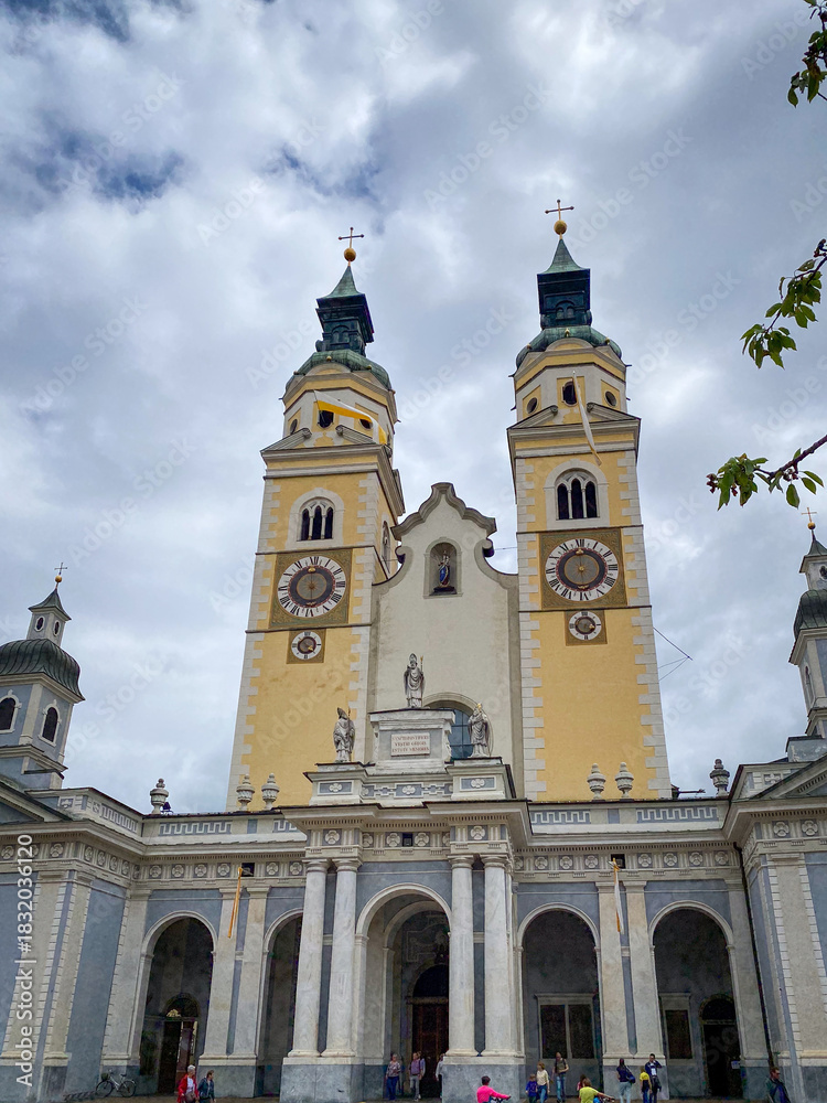 Obraz premium Twin towers of Brixen Cathedral in South Tyrol, Italy, yellow facades with copper spires and clock faces under a sky with clouds