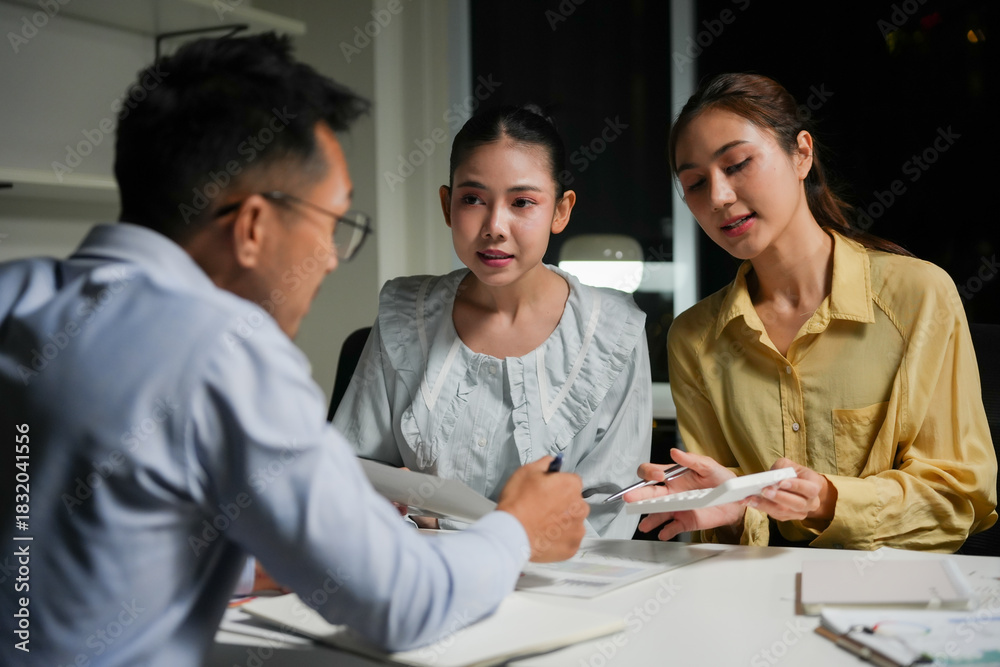 © Crystal - Young Asian businesspeople discussing project details and calculating figures while working late in a dark modern office
