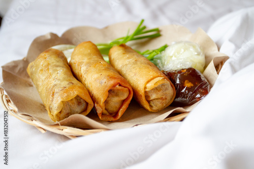 Three deep-fried Lumpia Semarang spring rolls sit on brown paper in a woven basket, served with fresh Allium fistulosum, pickled cucumber in plastic, and thick dipping sauce on a white surface.