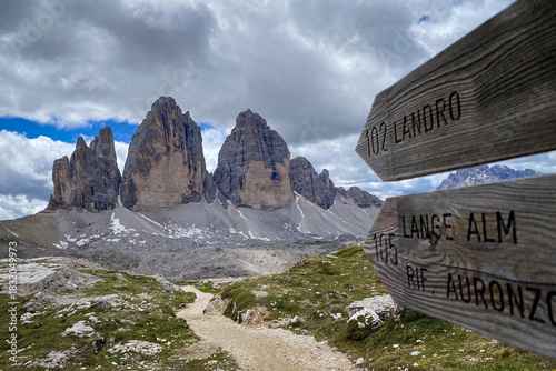 Wooden wayfinding signs for Landro, Lange Alm and Rifugio Auronzo on the Tre Cime loop; rocky path toward the three towers under a partly cloudy sky in Italy