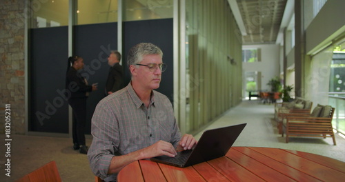 Middle aged man sitting at round table in office space, momentarily pensive and worried, then resuming work on laptop with focused determination despite visible concern