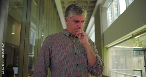 Middle aged man standing in office hallway with hand near mouth looking down anxious expression conveying worry stress and preoccupation in professional setting