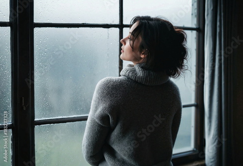 A woman stands by a window, gazing outside. She wears a gray turtleneck sweater and has her hair loosely tied up. The window is covered in raindrops, creating a misty background. 