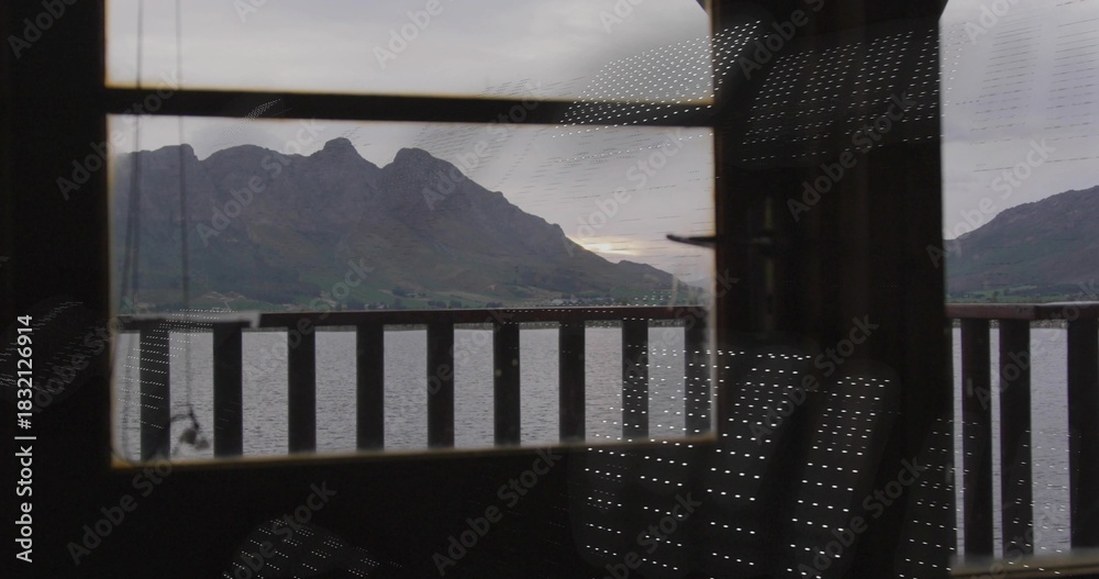 Naklejka premium Framing jagged mountain ridge and calm lake through window in dim cabin, with railing reflections
