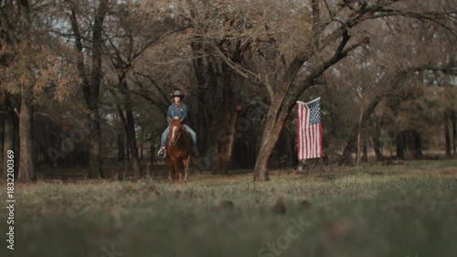 Cowgirl Woman With Braid and Scarf Wearing Cowboy Hat and Denim Jacket Riding Brown Horse With American Flag and Tree in Field in the Background