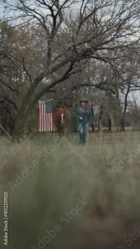 Cowgirl Woman With Braid and Scarf Wearing Cowboy Hat and Denim Jacket Walking With Brown Horse With American Flag and Tree in Field in the Background