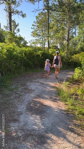 A woman and child hold hands while walking along a sandy path through green pines. Warm evening light creates a peaceful family nature outing.