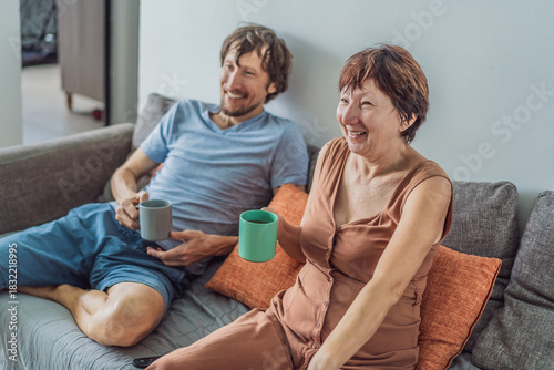 Older woman and her adult son relaxing together on the sofa while watching a comedy on television, enjoying a warm family moment at home. Entertainment, bonding and cozy lifestyle concept