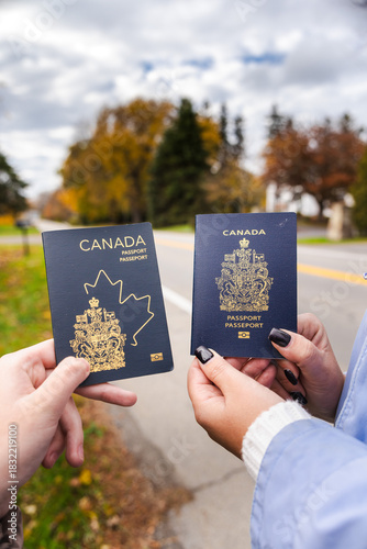 Two people outdoors holding Canadian passports side by side, showing the covers clearly against an autumn background near a road.