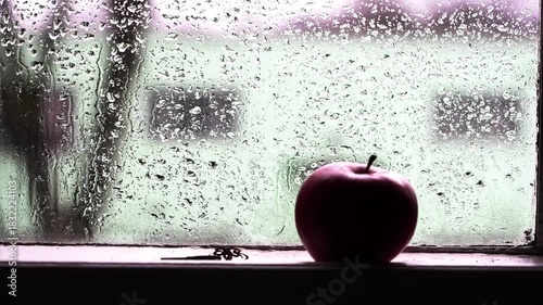 Raindrops create a hazy backdrop on the window while an apple rests on the sill. The soft glow adds a cozy ambiance to the rainy day setting.