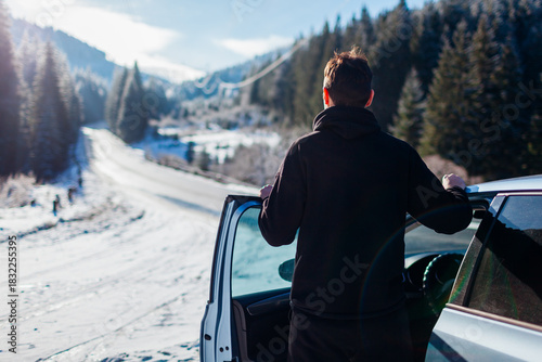 Back view of man admiring snowy mountains walking out of car looking at winter landscape. Driver traveling by auto