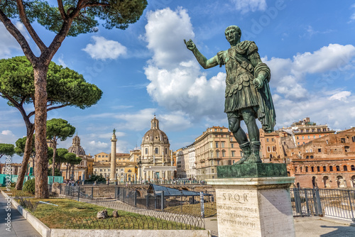 Statue of Julius Caesar at the Imperial Forums in Rome, Italy, with Trajan’s Column, historic churches, and ancient ruins in the background under a bright blue sky.