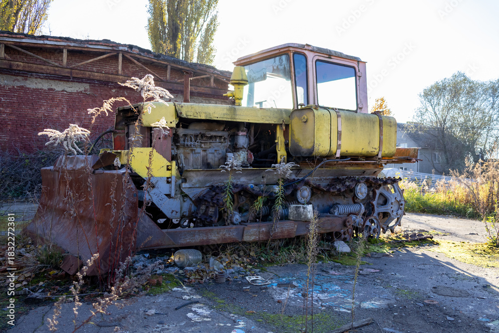 Fototapeta premium A yellow bulldozer sits rusting in a derelict industrial area, surrounded by overgrown weeds and remnants of an old building in the morning sun