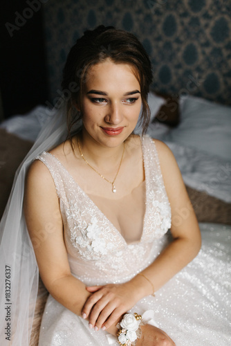 Portrait of a bride sitting indoors in her wedding dress. She looks at the camera with a calm, confident expression. Soft light highlights the lace details, veil, and floral bracelet, creating 