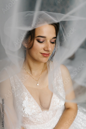 Bride smiling softly as her veil flows around her, creating a dreamy and romantic atmosphere. The close-up portrait highlights her makeup, lace wedding dress, and elegant jewelry in gentle natural 