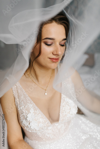 Bride smiling softly as her veil flows around her, creating a dreamy and romantic atmosphere. The close-up portrait highlights her makeup, lace wedding dress, and elegant jewelry in gentle natural 