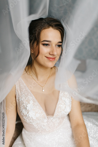 Bride smiling softly as her veil flows around her, creating a dreamy and romantic atmosphere. The close-up portrait highlights her makeup, lace wedding dress, and elegant jewelry in gentle natural 