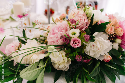 A delicate floral arrangement of pink and white peonies with small flowers decorates a festive table. Soft colors and greenery create a romantic, warm, and elegant atmosphere.
