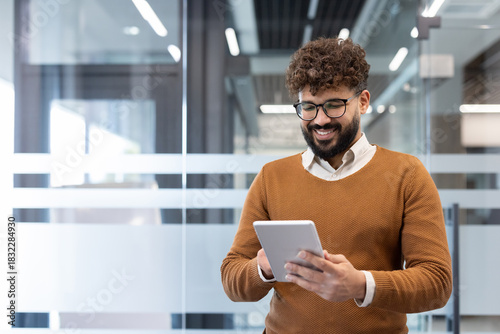 Young professional man in a contemporary office smiling as he uses a tablet to review business data and communicate online, reflecting tech-savvy, productive work life