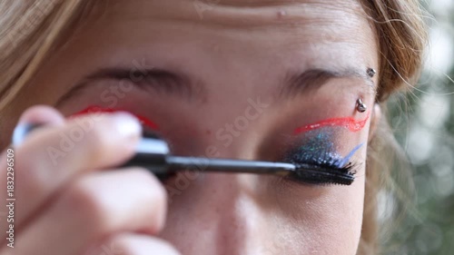 Caucasian girl applying mascara to her eyelashes, has an eyebrow piercing and brown eyes.