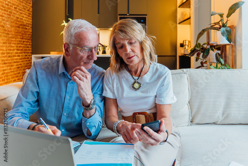 Elderly couple on couch seriously reviewing household budget and bills, using laptop and smartphone for online banking and payments while organizing paperwork at home