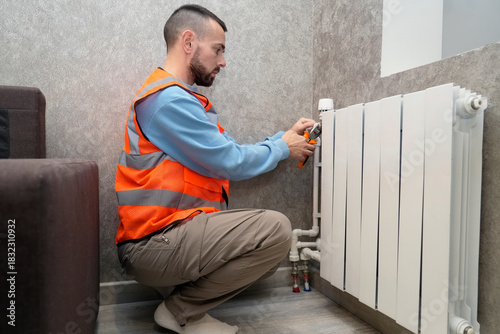 Worker specialist spends an hour repairing a radiator in a stylish living room. He wears a safety vest and focuses on fixing the heating system near the wall