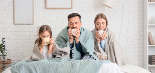 Cute little girl and her parents with warm plaids drinking hot tea in bedroom