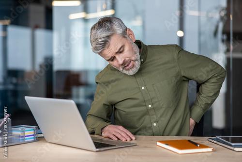 Mature man in an office slumped at his laptop, wincing and rubbing his lower back from strain and poor posture during a long sedentary workday, showing chronic discomfort