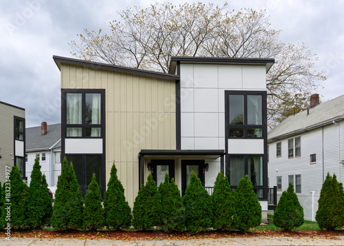 Modern two-story duplex with bold geometric facade and evergreen hedge in Brighton, Boston, Massachusetts, USA
