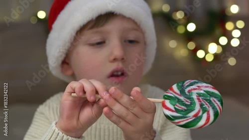 Joyful child enjoying colorful holiday lollipop with warm Christmas lights glowing in soft bokeh behind, creating cozy Christmas mood, magical seasonal moments. Cute boy looks at and twirls lollipop.