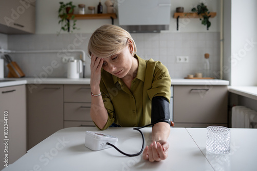 Middle aged woman with fatigue and headache feeling sick and measures blood pressure level with tonometer. Female showing signs of low blood pressure and loss of energy sitting at table in kitchen.