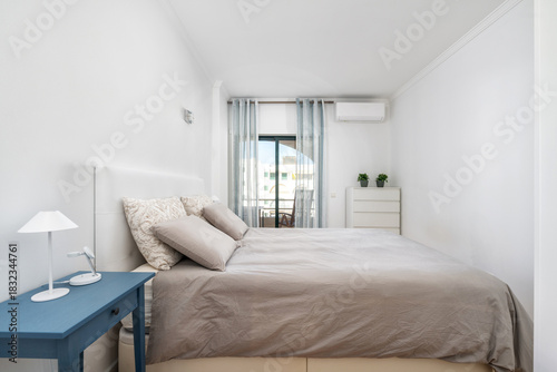 This bedroom features a large bed with soft linens, a small blue nightstand, and light curtains letting in natural light. A minimalist design enhances relaxation