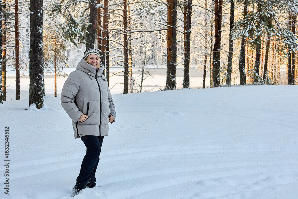 Fototapeta premium Winter walk through snowy forest, warmly dressed middle aged woman poses to someone against small frozen lake.