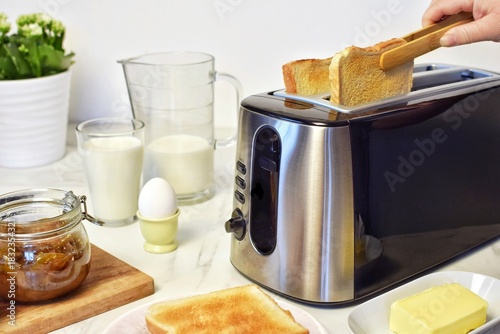 On the table is a modern toaster with slices of bread, butter, milk, and berries for breakfast. A woman removes a slice of bread from the toaster with tongs.