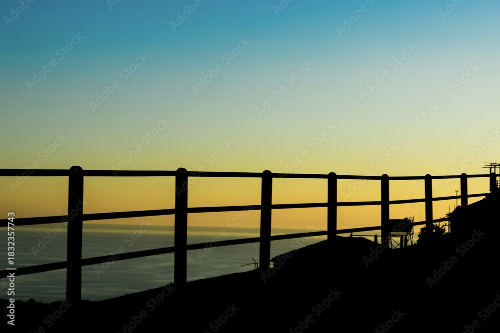 Fototapeta premium Silhouette of mountain road railing above sea at sunset, dramatic coastal travel landscape with vivid sky