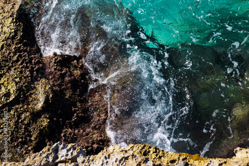 Top view of turquoise transparent wave with white foam hitting rocky seashore on a bright sunny day