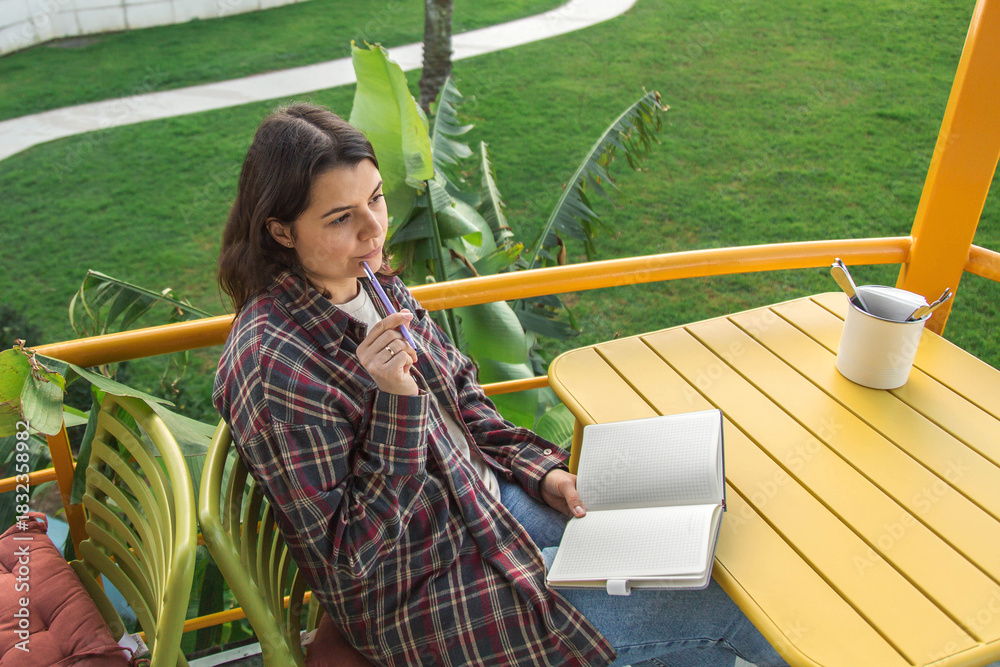 Obraz premium A woman sits on an outdoor terrace with an open notebook, thoughtfully reflecting on her goals and planning her day while enjoying the calm atmosphere, fresh air, and peaceful greenery around her.