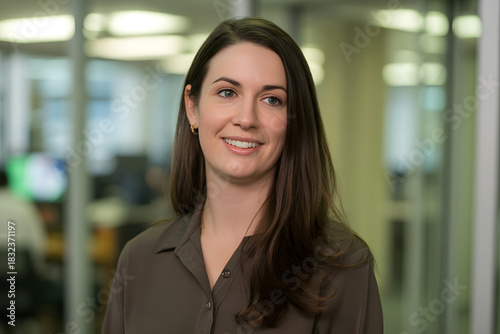 Free Download Professional headshot of a woman with long dark brown hair in a brown collared shirt, smiling warmly in a softly blurred office setting with glass partitions. - GFXLoad