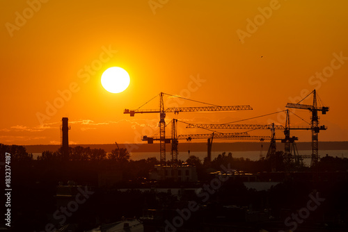 Silhouettes of multiple construction cranes and industrial structures are set against a vibrant orange sunset sky over Tallinn, with the sea visible in the distance