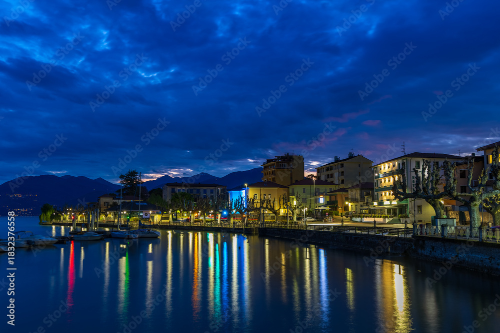 Naklejka premium Night view of the Porto Valtravaglia waterfront, featuring the brightly illuminated buildings reflecting on the calm lake surface beneath a dark, cloudy blue sky