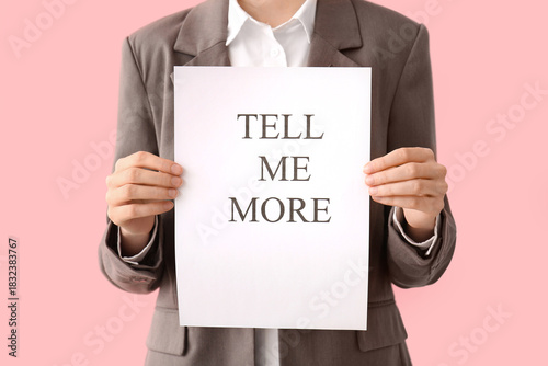 Businesswoman holding paper with text TELL ME MORE on pink background, closeup. Gossip concept