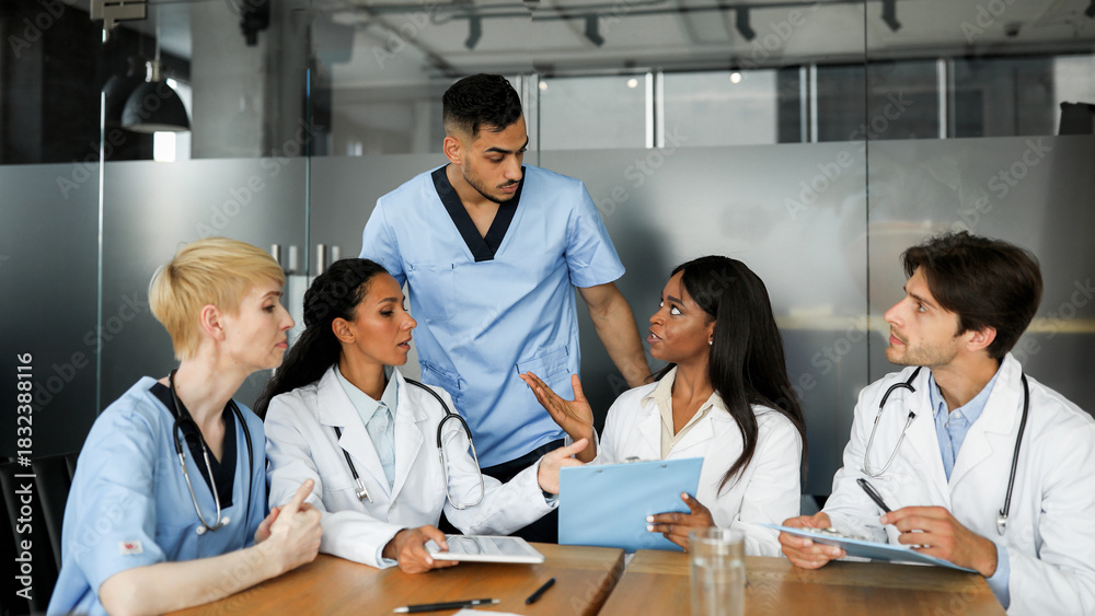 Fototapeta premium Concentrated multiracial group of doctors males and females in workwear having consultation, sitting at cabinet at hospital, discussing ways of treatment, panorama with copy space