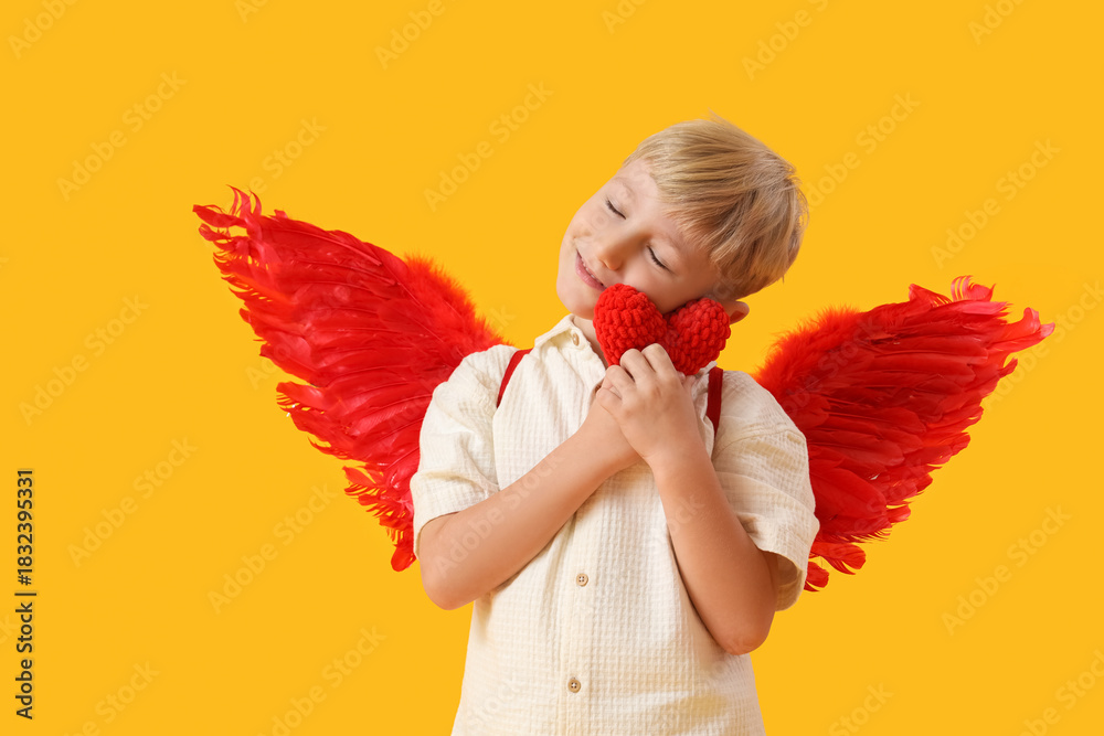 Obraz premium Cute little happy boy dressed as cupid with knitted heart toy on yellow background. Valentine's Day celebration