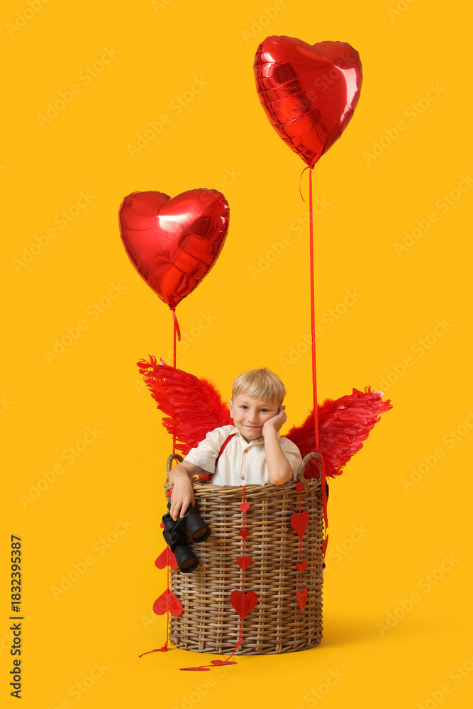 Fototapeta premium Cute little happy boy dressed as cupid in wicker basket with heart-shaped balloons and binoculars on yellow background. Valentine's Day celebration