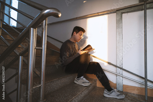 A young Asian man of Chinese descent is holding a book and sitting on the steps of an outdoor fire escape staircase in the late afternoon sunlight.