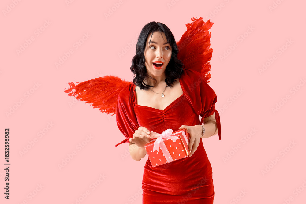 Fototapeta premium Beautiful young shocked woman dressed as cupid with gift box on pink background. Valentine's Day celebration