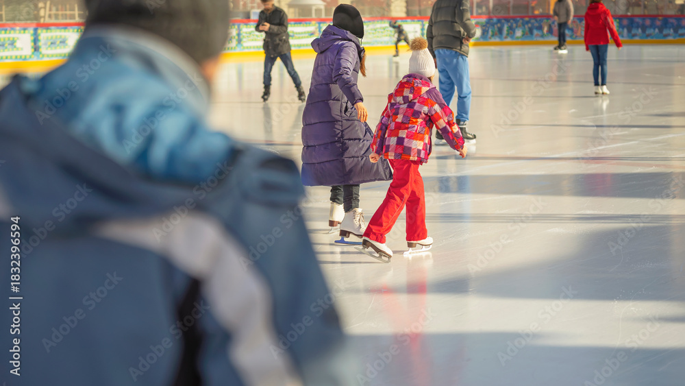Fototapeta premium People ice skating on open-air rink during sunny winter day, wearing colorful jackets and warm clothes. Concept of active winter leisure, joyful family ice skating experience