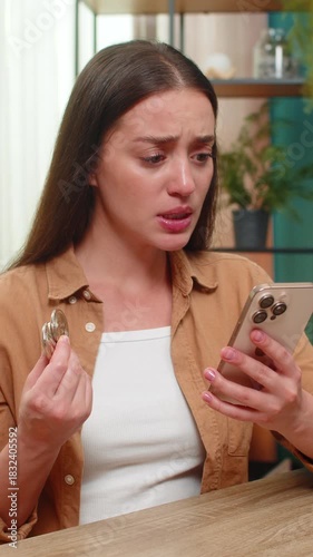 Young woman at home table watches smartphone graph as bitcoin price declines fast anxiety climbs high. Caucasian girl bites lip sensing financial loss, searches for hope while news continues to update